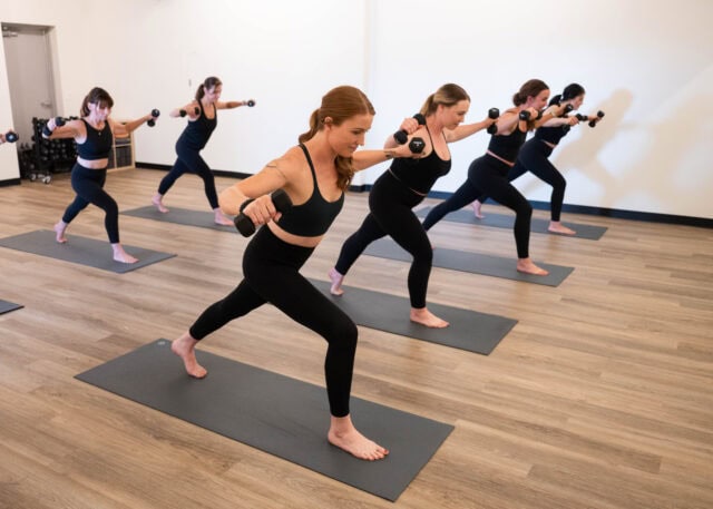 A group of people in exercise attire perform a lunge with dumbbells on yoga mats in a studio.