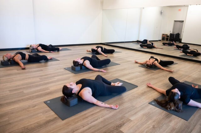 A group of people in a yoga studio lie on mats in a resting pose, eyes closed. The room has wooden floors and mirrored walls.